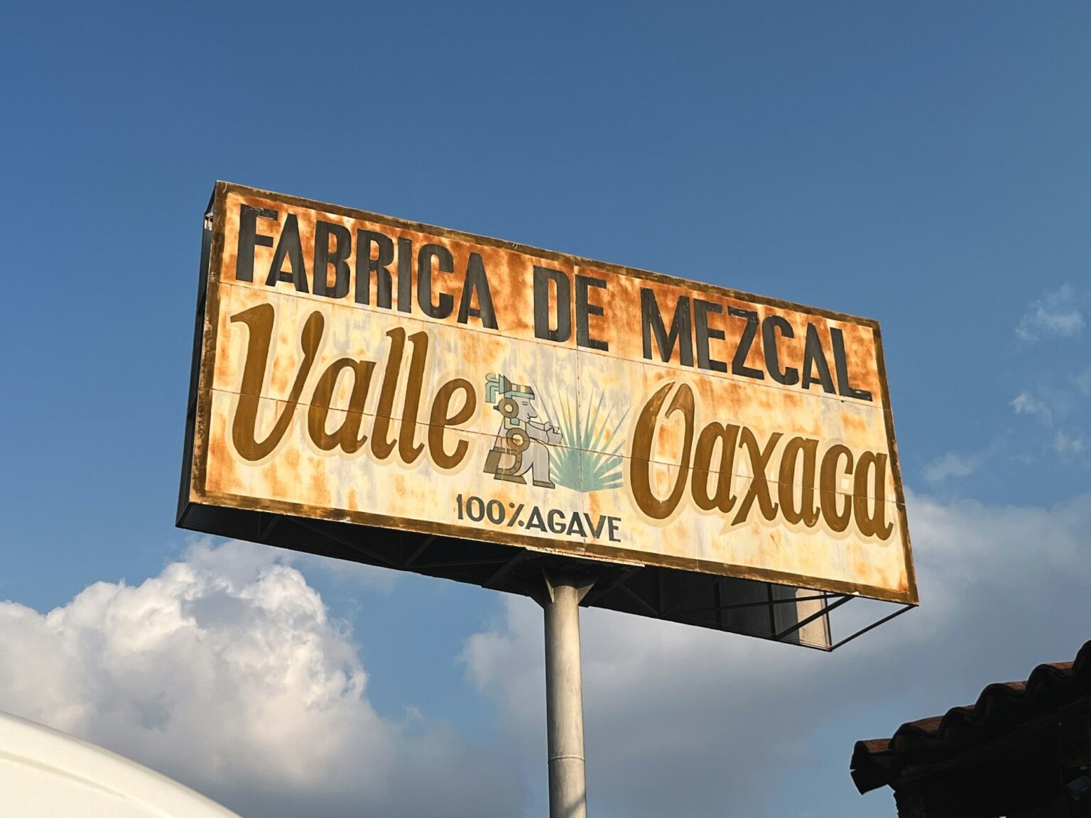 A weathered roadside sign for a mezcal factory in the Valley of Oaxaca against a blue sky.